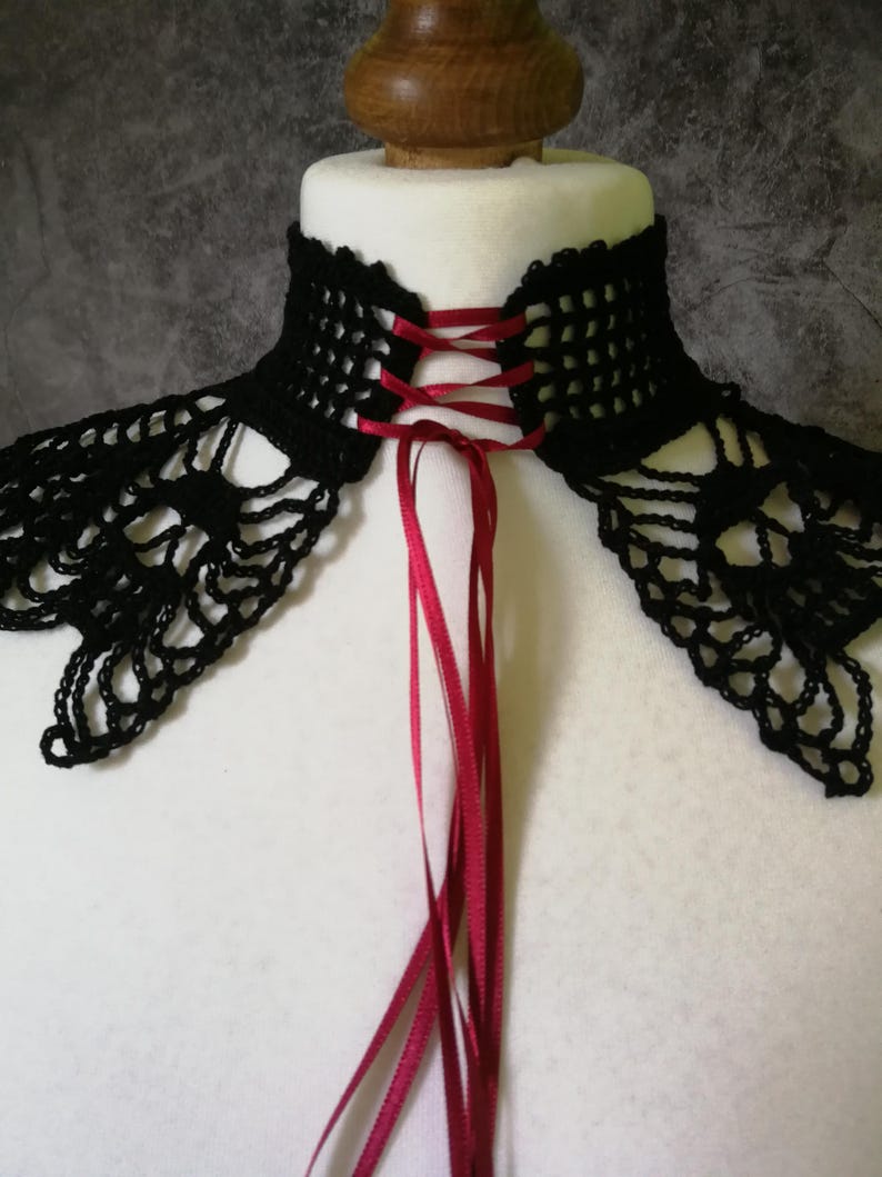 Puede incluir: Un cuello negro de ganchillo con un delicado dise&ntilde;o calado. El cuello presenta una cinta roja que se entrelaza en la parte posterior, creando un detalle tipo cors&eacute;. El cuello se muestra sobre un busto de maniqu&iacute; blanco.
