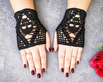 Può includere: Guanti senza dita all'uncinetto neri con un design traforato. I guanti presentano un motivo dettagliato e sono indossati su mani con smalto rosso scuro. È visibile un anello d'oro.
