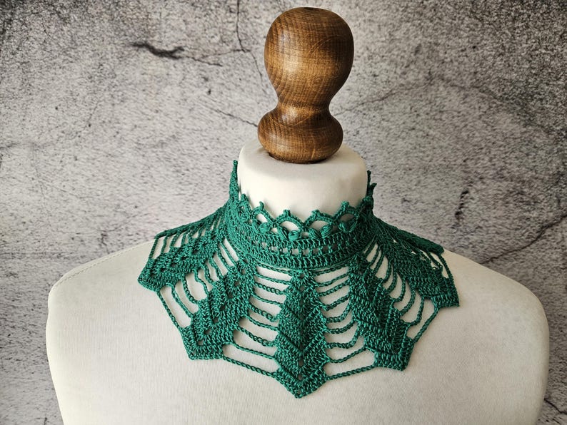 May include: A handmade, emerald green crochet collar. The collar is displayed on a white mannequin bust with a wooden neck form. The intricate openwork design features a scalloped edge and a geometric pattern.