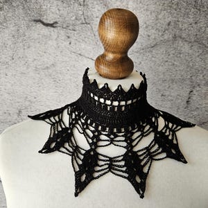 Op de afbeelding: Zwarte gehaakte kraag met een spinnenwebontwerp, weergegeven op een witte mannequin buste. De kraag heeft een geschulpte rand en een gedetailleerd, open patroon. De achtergrond is een getextureerde grijze muur.