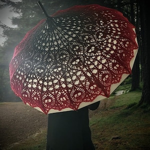 Puede incluir: Un paraguas decorativo con un diseño calado único. La cubierta presenta un degradado de color, pasando del rojo oscuro al negro. El intrincado patrón se destaca sobre un fondo blanco, creando un contraste visual llamativo. Un accesorio elegante.