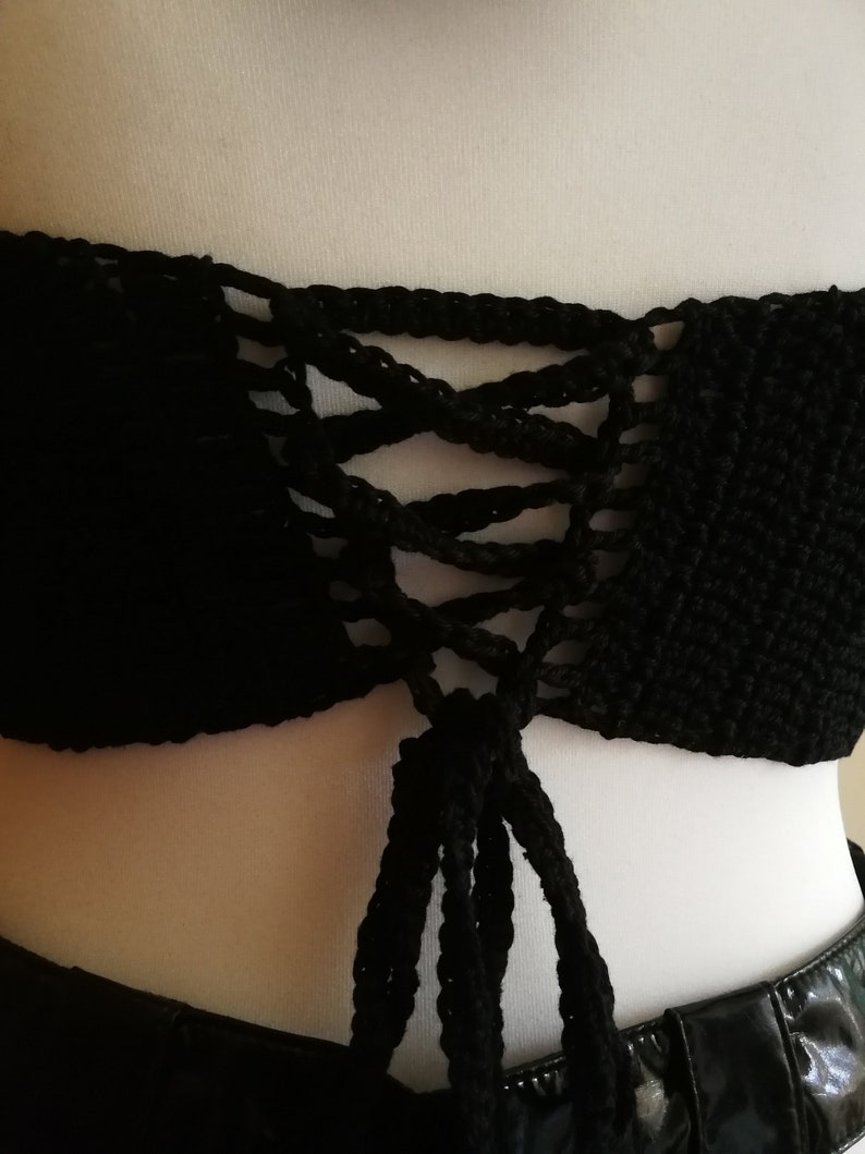 May include: Black crochet crop top with a lace-up front. The top is made of a solid black yarn and features a detailed, open-weave design. The lace-up detail adds a decorative element to the top.