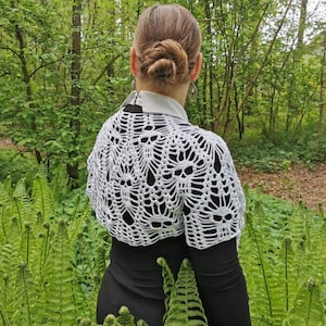 May include: A white crocheted shawl with a skull pattern. The shawl is worn over a black dress and the person is standing in a woodland setting.