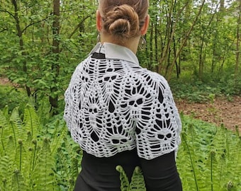 May include: A white crocheted shawl with a skull pattern. The shawl is worn over a black dress and the person is standing in a forest.