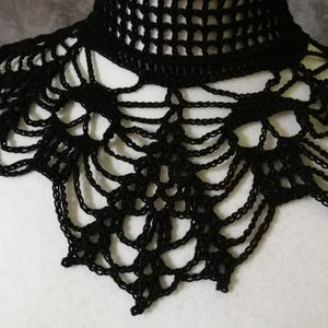 Puede incluir: Un cuello negro de ganchillo con un escote alto y calado y un dise&ntilde;o decorativo en forma de telara&ntilde;a. El cuello est&aacute; hecho de un material suave y flexible y probablemente se usa como accesorio de moda.