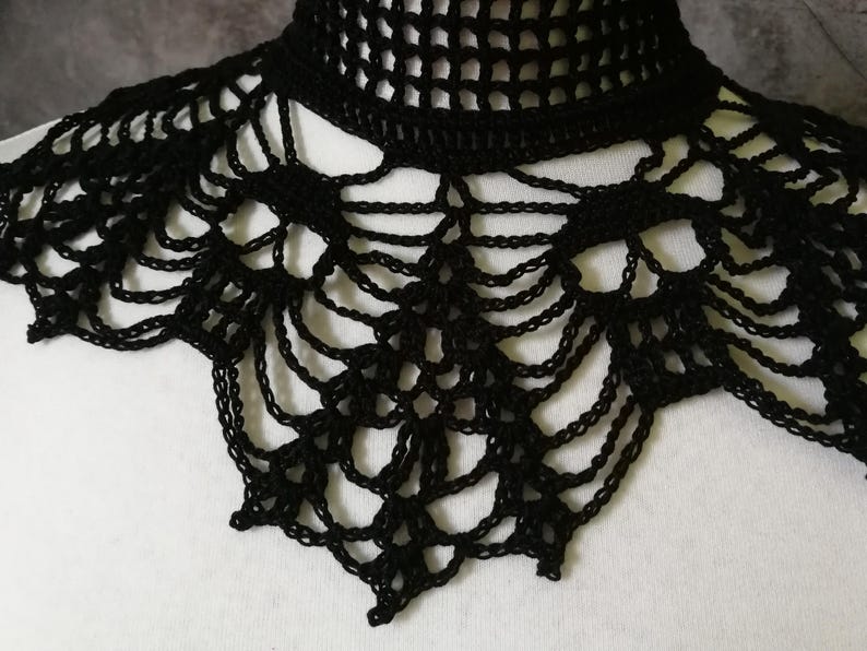Puede incluir: Un cuello negro de ganchillo con un escote alto y calado y un dise&ntilde;o decorativo en forma de telara&ntilde;a. El cuello est&aacute; hecho de un material suave y flexible y probablemente se usa como accesorio de moda.