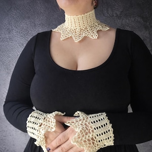 May include: Cream crochet collar and wrist cuffs. The high-necked collar has a scalloped edge, and the cuffs feature an open-work design with a similar scalloped pattern. The items are worn with a black long-sleeved top.