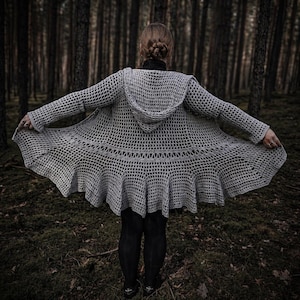 May include: A light gray, open-weave crochet hooded cardigan with a flared, ruffled hem. The long-sleeved garment is displayed outdoors, with the wearer's back to the camera, arms outstretched. The background is a dark forest.