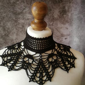 Puede incluir: Un collar negro de ganchillo con un dise&ntilde;o delicado en forma de telara&ntilde;a. El collar se exhibe sobre un busto de maniqu&iacute; blanco, mostrando su intrincado patr&oacute;n calado. El fondo es gris moteado.