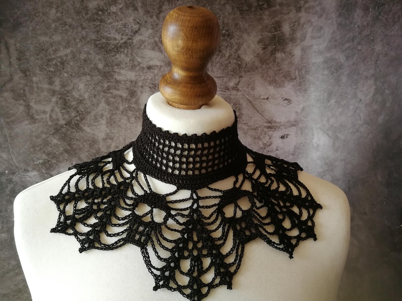 Puede incluir: Un collar negro de ganchillo con un dise&ntilde;o delicado en forma de telara&ntilde;a. El collar se exhibe sobre un busto de maniqu&iacute; blanco, mostrando su intrincado patr&oacute;n calado. El fondo es gris moteado.