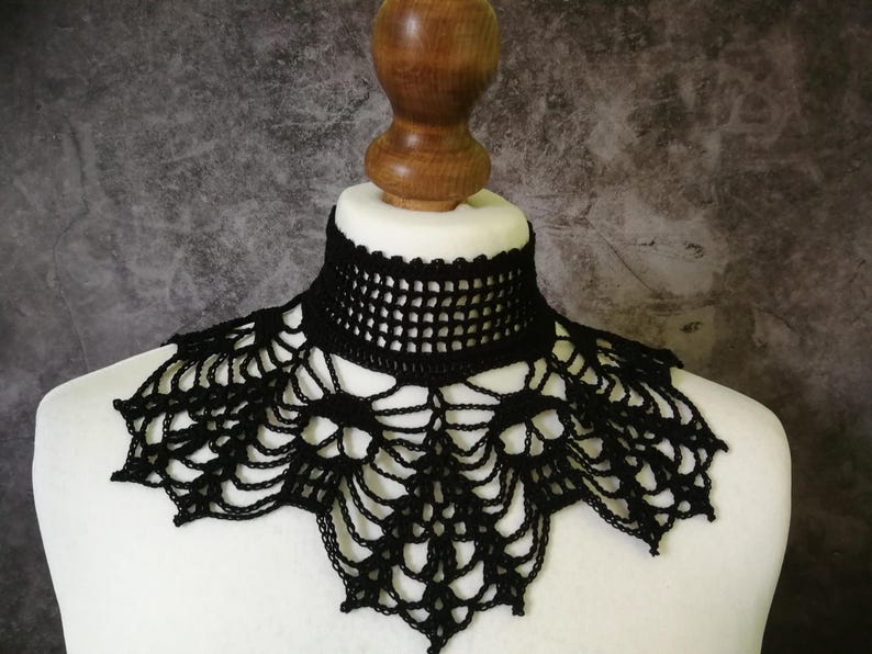 Puede incluir: Collar negro de ganchillo, exhibido en un busto de maniqu&iacute; blanco. El collar presenta un dise&ntilde;o detallado de tejido abierto alrededor del cuello y un patr&oacute;n decorativo de encaje que se extiende por el pecho. El fondo es gris moteado.