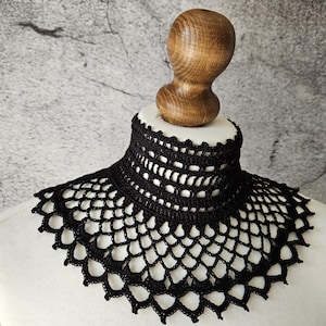 May include: A black, crocheted collar displayed on a white mannequin bust. The collar features an open, lacy design with a high neckline and scalloped edges. The bust is topped with a wooden head form. The background is a mottled gray.