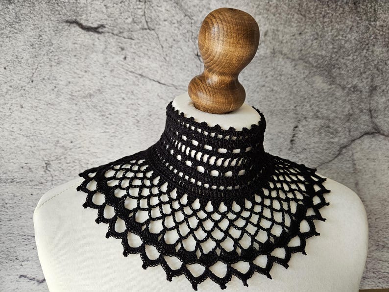 May include: A black, crocheted collar displayed on a white mannequin bust. The collar features an open, lacy design with a high neckline and scalloped edges. The bust is topped with a wooden head form. The background is a mottled gray.