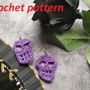 Puede incluir: Un par de pendientes de calavera de ganchillo morado con ganchos plateados. Los pendientes están sobre una superficie gris texturizada.