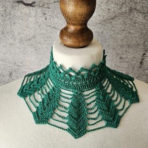 May include: A handmade, emerald green crochet collar. The collar features a delicate, openwork design with a scalloped edge. The collar is displayed on a white mannequin head, with a wooden stopper in the background.