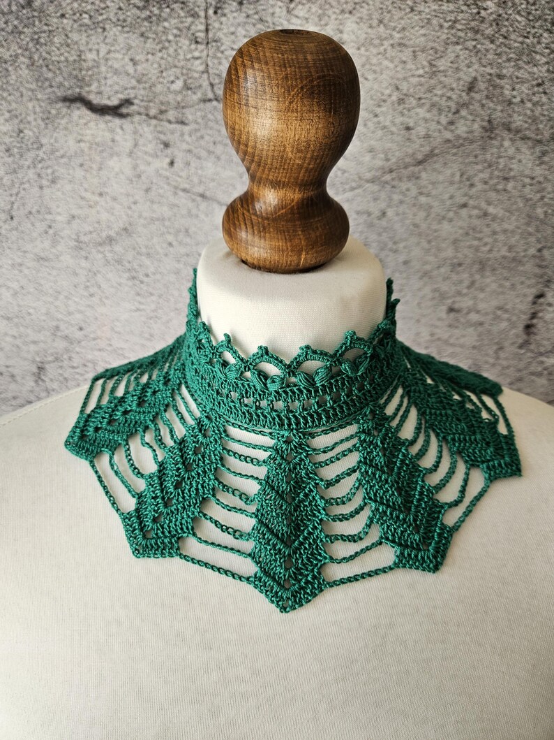 May include: A handmade, emerald green crochet collar. The collar features a delicate, openwork design with a scalloped edge. The collar is displayed on a white mannequin head, with a wooden stopper in the background.