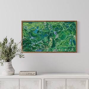 The vibrantly green and colorful Forest Park painting framed in oak on white wall in home interior setting above a simple wooden console with a book and a vase with greenery.