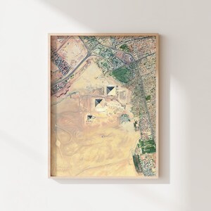 Op de afbeelding: Een ingelijste luchtfoto van de piramides van Gizeh in Egypte. De prent toont de piramides omgeven door woestijn en stedelijke gebieden, in een lichtgekleurde houten lijst. De afbeelding is een gedetailleerde, kleurrijke weergave van de oude site.