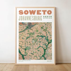 Op de afbeelding: Een ingelijste wandkunstprint met een gedetailleerde luchtfoto van Soweto, Johannesburg, Zuid-Afrika. De tekst SOWETO staat in grote perzikkleurige letters boven de woorden JOHANNESBURG SOUTH AFRICA in het groen. De kaart toont stedelijke rasters.