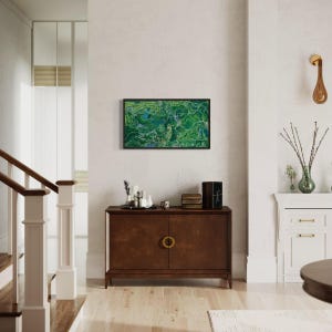 The vibrantly green and colorful Forest Park painting framed in walnut on wall in traditional home interior setting above a wooden console with modern, neutral accents and high-end finishes and light fixtures.