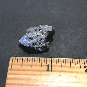 May include: A raw, uncut mineral specimen with a mix of dark gray, black, and light blue hues. The crystal formation has a rough, textured surface. A wooden ruler with inch markings is in the foreground.