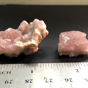 May include: Two raw, pink mineral specimens. The larger piece is a cluster of crystals, and the smaller is a cube-shaped stone. A ruler with inch measurements is in the foreground, indicating the size of the minerals.