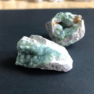 May include: Two raw mineral specimens featuring clusters of pale green, botryoidal crystals embedded within a rough, light gray rocky matrix. The crystalline formations exhibit a textured, rounded surface with a soft, translucent appearance.