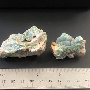 May include: Two raw mineral specimens with a ruler for scale. The minerals have a light green, bubbly texture on a tan and brown matrix. The ruler shows measurements in inches.