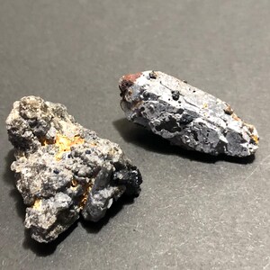 May include: Two rough, irregularly shaped rocks with a gray, black, and orange color palette. The rocks have a textured, granular appearance, suggesting a mineral or geological specimen. The background is a solid, dark gray surface.