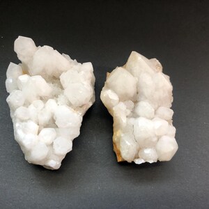 May include: Two clusters of white quartz crystals on a dark gray background. The crystals are clustered together in a natural formation.