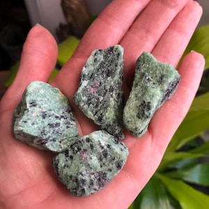 May include: Four rough, green and black, natural, tumbled, zultanite gemstones. The stones are in the palm of a hand.