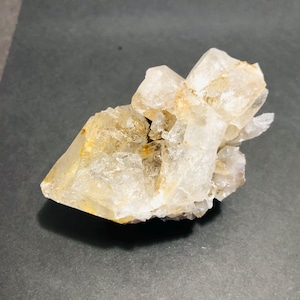 May include: A cluster of translucent quartz crystals with a pale yellow hue. The crystals have sharp edges and varying sizes, creating a textured appearance. The mineral formation sits on a dark gray surface.