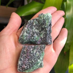 May include: Two rough, green and black, raw, uncut, natural, gemstone crystals. The crystals have a speckled appearance with hints of pink.