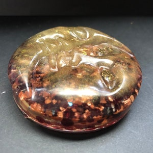 May include: A round, clear resin paperweight with embedded copper and gold flakes. The resin is translucent, allowing the flakes to be seen through the clear resin.