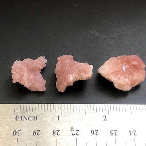 May include: Three raw, pink rhodochrosite mineral specimens. The stones are a light pink color and have a rough, textured surface. A ruler is placed below the stones to show their size.