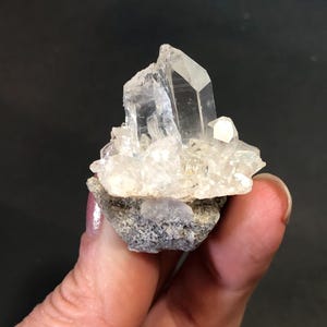 May include: A clear quartz crystal cluster with multiple points, resting on a base of gray and white rock. The quartz crystals are transparent and reflect light, showcasing their natural formations. The cluster is held in a hand.