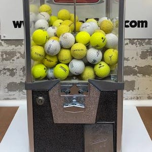 May include: A vintage gumball machine filled with golf balls. The machine has a clear glass front, a silver and black metal frame, and a coin slot. The golf balls are yellow and white.