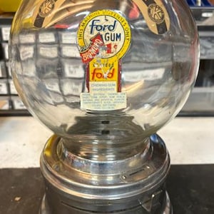 May include: A vintage glass gumball machine with a metallic base and a spherical clear container. The glass features a label reading "Underprivileged Children" and "Ford Gum" with a one-cent price indicator. The base includes a coin slot and lever.