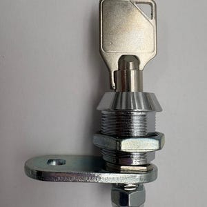 May include: A silver-colored cam lock with a key inserted. The lock has a cylindrical body, a mounting bracket, and a nut. The key is rectangular with a cut-out design. The lock is designed for security purposes.