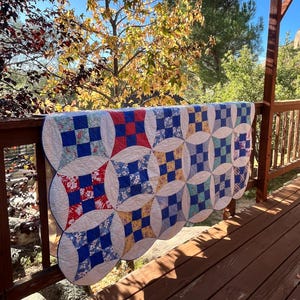 May include: A handmade quilt with a repeating pattern of blue, red, and yellow squares set within white circles. The quilt is draped over a wooden railing on a sunny day, with trees and a blue sky in the background.
