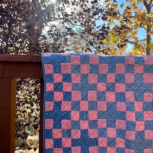 May include: A handmade quilt with a checkerboard pattern of pink and blue squares. The quilt is hanging on a wooden fence outdoors, with trees and sunlight in the background. The quilt is a traditional style, likely made of cotton.