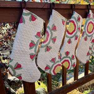 May include: Four quilted Christmas stockings with floral designs hang from a wooden railing. The stockings are white with red, green, and yellow floral patterns. The background shows fall foliage.