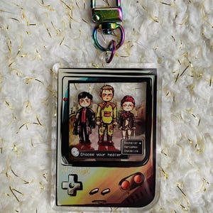 Puede incluir: Un llavero acrílico transparente con un diseño de videojuego. Presenta tres personajes y el texto "Choose your healer". El llavero tiene un cierre iridiscente colorido.