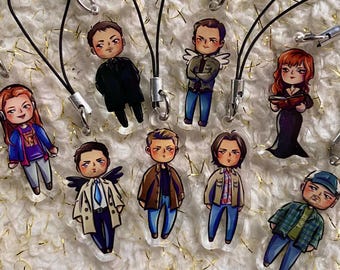 May include: A selection of cartoon character charms with black lanyards. The charms depict various characters in different outfits, including a black coat, a tan jacket, and a blue plaid shirt. The background is a white, textured surface with gold accents.