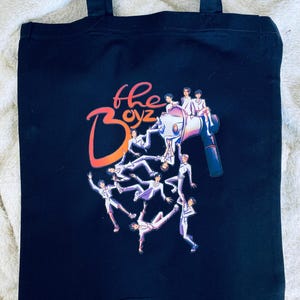 May include: Black tote bag featuring the text "the Boyz" in orange and white, with an illustration of several figures in white suits and a megaphone. The bag has two black handles.