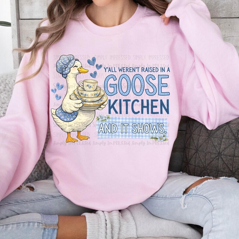 May include: A light pink sweatshirt featuring a cartoon goose wearing a bonnet and apron, holding a stack of teacups and saucers. The text reads "Y'all weren't raised in a Goose Kitchen and it shows."