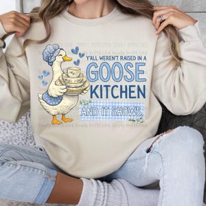 May include: Beige sweatshirt with a goose graphic holding dishes and the text "Y'all weren't raised in a Goose Kitchen and it shows." The goose wears a blue bonnet and apron. The text is in blue.