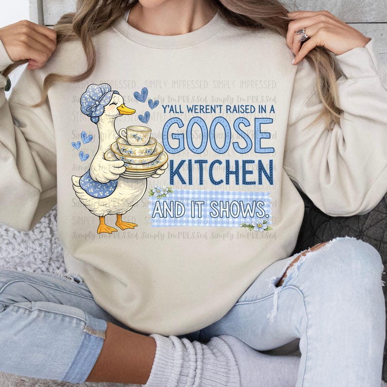 May include: Beige sweatshirt with a goose graphic holding dishes and the text "Y'all weren't raised in a Goose Kitchen and it shows." The goose wears a blue bonnet and apron. The text is in blue.
