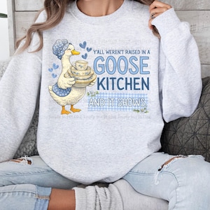 May include: A light gray sweatshirt with a goose graphic holding a stack of teacups and plates. The text "Y'all weren't raised in a GOOSE KITCHEN AND IT SHOWS" is in blue. The goose wears a blue bonnet and apron.