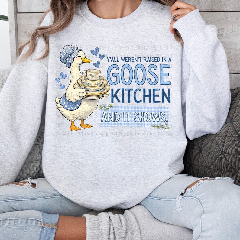 May include: A light gray sweatshirt with a goose graphic holding a stack of teacups and plates. The text "Y'all weren't raised in a GOOSE KITCHEN AND IT SHOWS" is in blue. The goose wears a blue bonnet and apron.